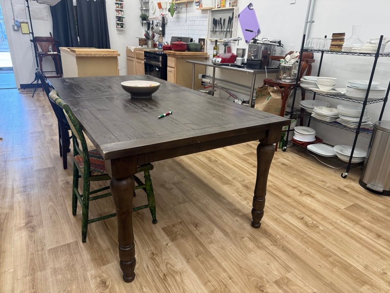 Large wooden dining table with black chairs in a studio or workshop space with shelving and storage units in background